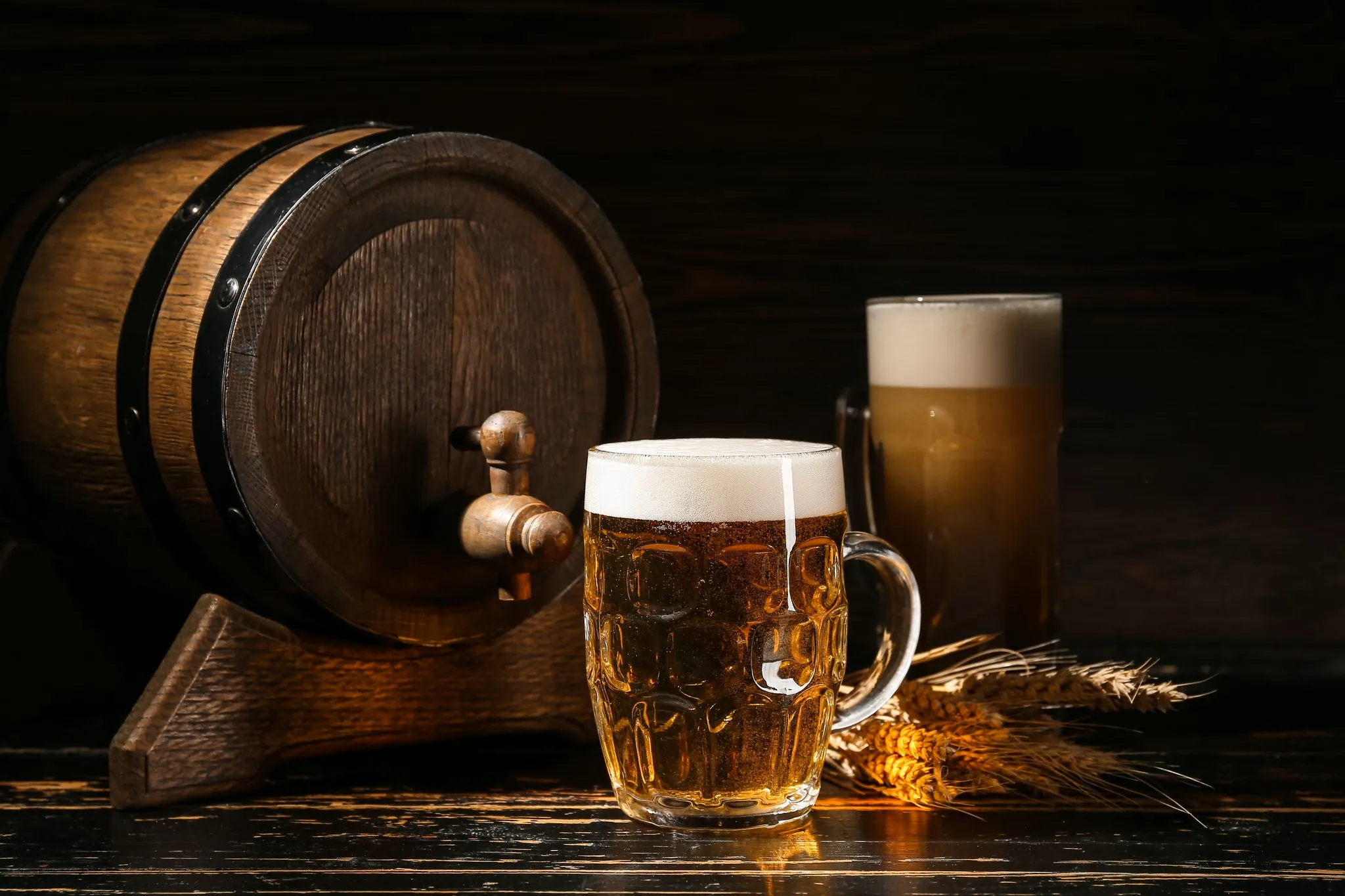Bierkrug mit Schaumkrone neben einem Holzfass mit Zapfhahn und Getreideähren auf einem dunklen Holztisch