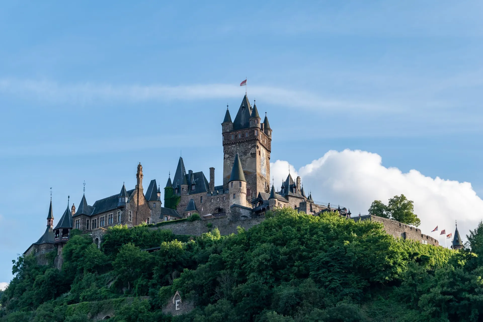 Die Reichsburg Cochem auf bewaldetem Burgberg über der Mosel, im Hintergrund blauer Himmel mit Wolken