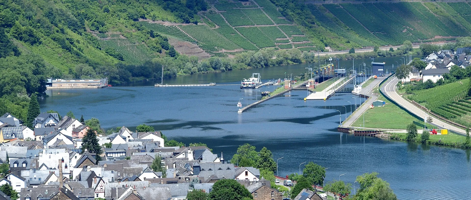 Staustufe Fankel an der Mosel mit Wehr und Schleusenkammer, davor ein Moseldorf, dahinter Weinberge in Steillage